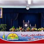 سالن مراسمات مزیّن شده با پرده آبی، سکو و میز رسمی، تزیینات گل رنگارنگ، و بنر مدرسه برای برگزاری مراسم یا اجتماع رسمی