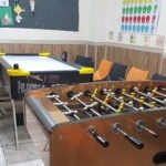 کلاس درس مدرسه‌ای با میزهای خاکستری، صندلی‌های نارنجی، تخته سیاه و بازی فوسبال