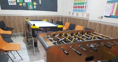 کلاس درس مدرسه‌ای با میزهای خاکستری، صندلی‌های نارنجی، تخته سیاه و بازی فوسبال