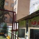 نمای حیاط مدرسه با ساختمان آجری، درب سرخ و حفاظ فلزی سبز