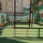 نمایی از حیاط مدرسه با دروازه‌های فوتبال و تجهیزات ورزشی بر روی زمین سبز، با پرچم ایران در پس‌زمینه