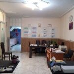 فضای داخلی دفتر اداری مدرسه با میز‌های کاری، صندلی‌ها، کابینت‌های ذخیره‌سازی، تجهیزات اداری و نورافکنی