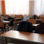 اتاق کلاس درس با میز‌های کاری قهوه‌ای، صندلی‌های دانش‌آموز و پنجره‌های بزرگ با پرده‌های نارنجی و سفید
