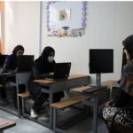 گروهی از دانش‌آموزان با ماسک و مدرس در کلاس کامپیوتر، دانش‌آموزان با لپ‌تاپ روی میزهای آموزشی، نمای کلاس آموزش فناوری اطلاعات