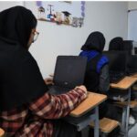 دختران دانش‌آموز در حال کار با لپ‌تاپ در اتاق رایانه‌ای مدرسه، با یکی از آن‌ها ایستاده در کنار دسک‌های چوبی