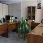 اتاق کتابخانه با قفسه‌های شیشه‌ای پر از کتاب‌ها، میز‌های چوبی، کمپیوتر و صندلی‌های اداری