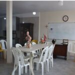 دانش‌آموزی ایستاده در کلاس درس درحال انجام ارائه یا فعالیت درسی، با صندلی‌های زرد و سفید دور میز، تخته سفید و ساعت دیواری