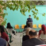 مردی با ماسک در حال سخنرانی جلوی گروه افراد نشسته در حیاط مدرسه با دیوار سبز آبی