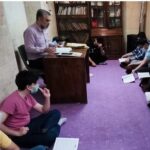 یک معلم در حال تدریس درس در کنار دانش‌آموزانی که روی فرش بنفش‌رنگ نشسته‌اند و کتاب و دفترچه در دست‌ها دارند، با کتاب‌های فراوان روی شلف‌های چوبی در پس‌زمینه