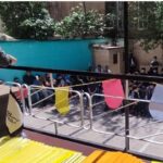 سخنران ایستاده در محراب سخنرانی در حال صحبت به مجمع دانش‌آموزان نشسته در صندلی‌های رنگی در فضای باز (حیاط یا سالن) مدرسه