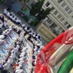 تصویری از یک مراسم یا رویداد دسته‌جمعی در حیاط مدرسه با حضور تعداد زیادی دانش‌آموز و پرچم‌های سرخ و سبز