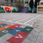 نمای حیاط مدرسه با کفپوش رنگی و الگوی بازی‌آموز، نقاشی‌های دیواری هنری، و فضایی باز برای بازی و فعالیت‌های فیزیکی دانش‌آموزان