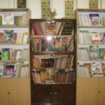 کتابخانه مدرسه با سه قفسه فلزی سفید و قهوه‌ای پر از کتاب‌های مختلف و مواد آموزشی، دارای دسته‌های متنوع و منظم