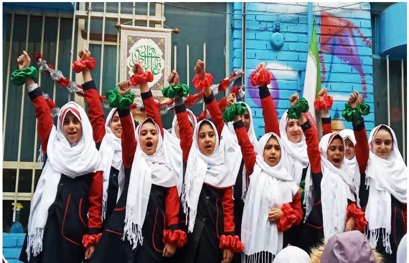 دانش‌آموزان دختر با لباس مدرسه و روسری سفید در حال برگزاری مراسم کاروان قرمز، با گل‌های قرمز در دست، در حیاط مدرسه