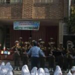 تصویری از مراسم رسمی با افراد بسیج/نیروی انتظامی در لباس نظامی ایستاده در تشکیل‌بندی سه‌ردیفی جلوی ورودی مدرسه با ساختمان آجری در پس‌زمینه
