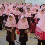 گروهی از دانش‌آموزان دختری با لباس مدرسه و روسری صورتی در حیاط مدرسه برای یک مراسم جمعی جمع شده‌اند