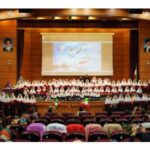 دانش‌آموزان مدرسه در لباس یکدست سفید، ایستاده بر روی صحنه‌ی سالن مراسم با دیوارهای چوبی و نورپردازی حرفه‌ای، در جریان مراسم تجلیل و اجتماع مدرسه