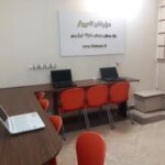 اتاق سایت کامپیوتر مدرسه با میز‌های تحریر، لپ‌تاپ‌ها و صندلی‌های قرمز
