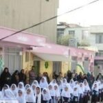 دانش‌آموزان دختری پوشیده به لباس مدرسه در یک مراسم اجتماعی در حیاط مدرسه با ساختمان‌های آجری پس‌زمینه ایستاده‌اند