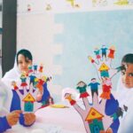 دو دانش‌آموز در کلاس درس در حال کار روی یک پروژه هنری و ساختار سه‌بعدی با قطعات رنگی و قالب‌های آموزشی