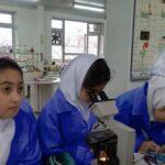 دانش‌آموزان دختر با لباس آزمایشگاه آبی در حال استفاده از میکروسکوپ و انجام آزمایش در آزمایشگاه مدرسه