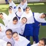گروهی از دانش‌آموزان دختر با لباس مدرسه و چادر آبی و سفید، در حیاط مدرسه ایستاده و به دوربین خیره شده‌اند