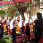 دانش‌آموزان مدرسه در حیاط برای یک مراسم یا تجمع جمعی، یکی از معلمان ایستاده در حال سخنرانی یا هدایت است، درخت بزرگی در پس‌زمینه