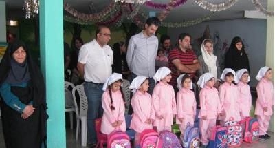 تصویری از مراسم و جشن مدرسه‌ای با حضور گروهی از دانش‌آموزان در لباس صورتی‌رنگ یکسان و تحت نظارت معلمین در سالن داخلی مدرسه با تزئینات و گل‌های مصنوعی در بالا