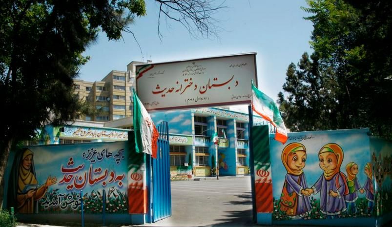 نمای بیرونی ورودی اصلی دبستان و دبیرستان هادی شریف با تابلوی نام مدرسه، دیوار رنگی آبی و نقاشی شاد کودکان، درهای آبی و پرچم‌های ایرانی