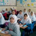 گروهی از دانش‌آموزان دختر با لباس یکپارچه و حجاب، نشسته به میزهای چوبی در کلاس درسی با دیوارهای تزئین‌شده با نقاشی و پوسترهای رنگین