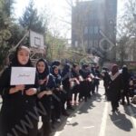 دانش‌آموزانی در لباس مدرسه در حیاط مدرسه حاضر در یک مراسم یا رویداد گروهی با برگه‌های کاغذی