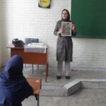 دانش‌آموزان در یک کلاس درس، یکی از آنها ایستاده و ارائه می‌دهد، تخته سیاه و میزهای درس در پس‌زمینه