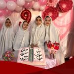 گروه دانش‌آموزانی با لباس سفید رسمی و حجاب سفید در حال پوز برای عکس جشن، با دکوراسیون بالون و گل صورتی، کیک مزیّن شده با متن فارسی و اعداد رنگین، برای جشن ادبی‌های مدرسه‌ای
