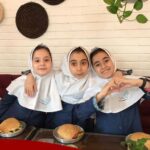 سه دانش‌آموزه با روسری و یونیفرم مدرسه در حال غذا خوری در یک رویداد، نشسته بر روی صندلی قرمز در اطراف میز غذا