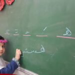 دانش‌آموزی در حال نوشتن حروف فارسی روی تخته سیاه کلاس درس، با تاج صورتی و روسری سفید