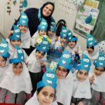 دانش‌آموزان و معلم در کلاس درس در حال انجام یک فعالیت هنری/خلاقانه با کاپ‌های رنگی آبی و سفید، در کنار تابلو سبز و پوسترهای کلاسی.