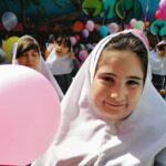 دانش‌آموزان دختر با روسری سفید و لباس مدرسه در حال شرکت در مراسمی رنگی، با بادکنک‌های صورتی و رنگین و تزئینات دیواری رنگارنگ