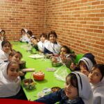 دانش‌آموزان در حال غذاخوری در سالن غذاخوری مدرسه با میز و صندلی‌های سبز و دیوار آجری