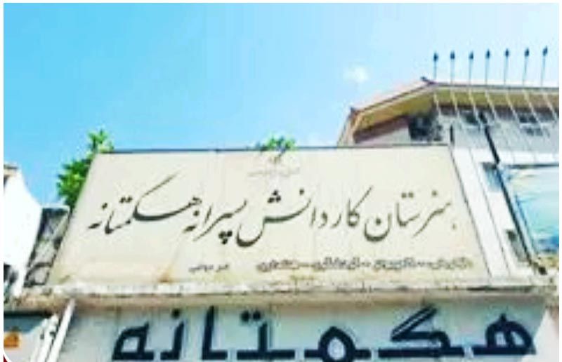 تابلوی نام‌گذاری ساختمان مدرسه هگمتانه به حروف فارسی بر روی دیوار سفید رنگ با آسمان آبی در پس‌زمینه