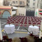 حیاط مدرسه با صندلی‌های قرمز و میز‌های تزئین شده برای برگزاری مراسم رسمی، پرچم‌های ایرانی و ستون‌های ساختمان