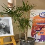 نمایشگاه هنری در فضای داخلی مدرسه با نمایش عکس‌های سیاه و سفید، گلدان سبز، و بنر پروژه‌ای موقت