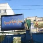 درب و سردر ورودی ساختمان مدرسه با تابلوی نام‌گذاری آبی رنگ و دیوار سفید