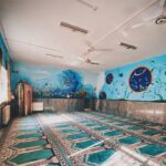 نمای داخلی نمازخانه مدرسه با قالیچه‌های نمازی آبی و سفید، دیوارهای آبی تزیین‌شده با نقوش هندسی و تصاویر مذهبی، و روشنایی سقفی