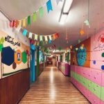 نمای داخلی راهرو مدرسه با دیوارهای صورتی و سبز، تزئین‌های فصلی رنگین، و درب‌های جمع‌کننده آثار دانش‌آموزان
