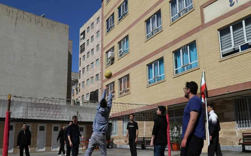 دانش‌آموزان در حیاط مدرسه در حال بازی والیبال تحت نظارت اساتید، با ساختمان آجری مدرسه در پس‌زمینه