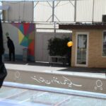 دانش‌آموزان در حال تعامل با میز تاچ تعاملی هوشمند در حیاط مدرسه، کنار نقاشی دیواری رنگین با طرح‌های هندسی