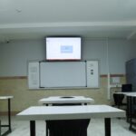 نمای عمومی از سایت کامپیوتری مدرسه با میزهای کار سفید، صندلی‌های سیاه، نمایشگر دیواری LCD، قفسه‌های ذخیره‌سازی آبی و تجهیزات شبکه
