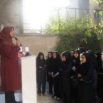 معلم یا مربی در حال سخنرانی به گروهی از دانش‌آموزان دختر با حجاب در حیاط مدرسه، در حالی که آنها با توجه گوش می‌دهند