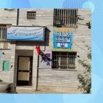 دیوار جانبی ساختمان مدرسه با درب ورودی، پنجره‌های توری‌دار، تابلوی شناسایی آبی و پرچم ایران
