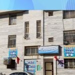 ساختمان سه طبقه‌ای سفید رنگ مدرسه ایران تکنیک با تابلوهای اطلاعاتی و پرچم ایران در نمای رو به جنوب